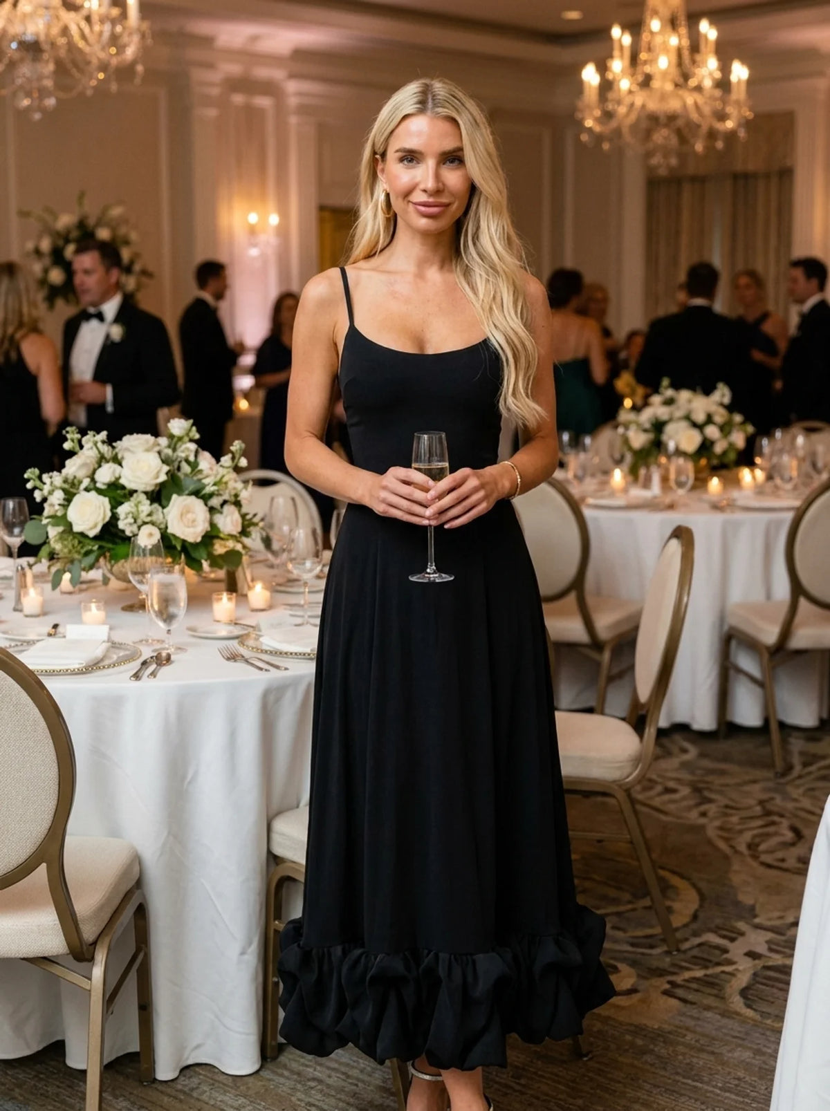 A blonde woman wearing a black spaghetti-strap maxi dress with a ruffled hem, holding a wine glass in a decorated event hall with tables and guests in the background.
