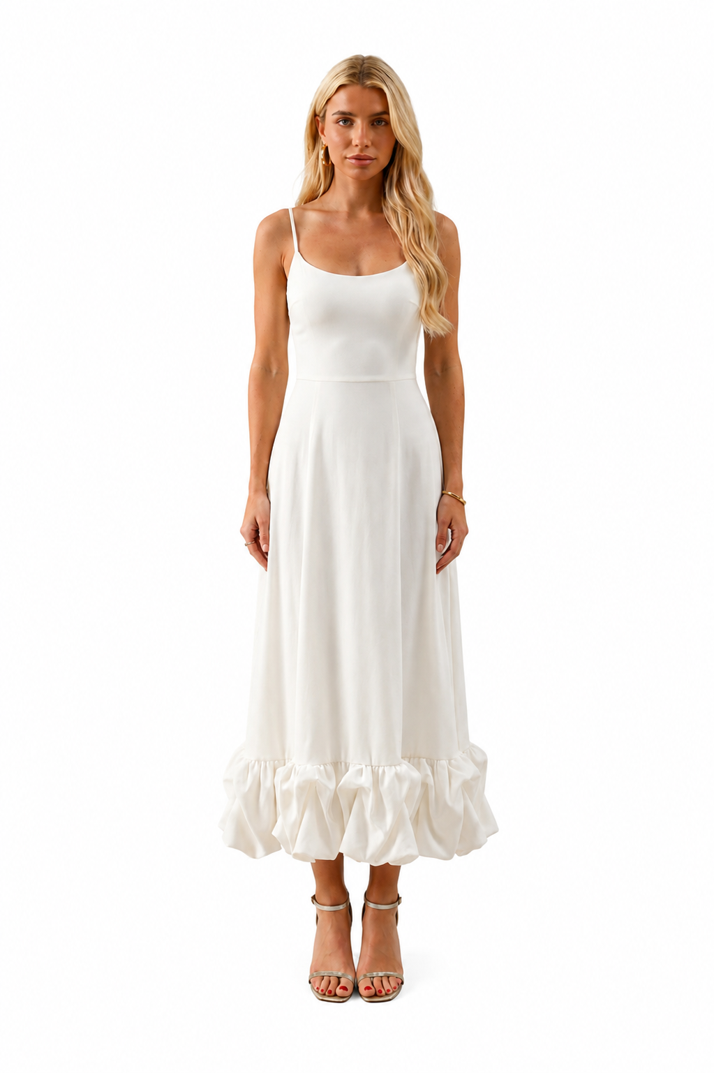A blonde model wearing a white spaghetti-strap maxi dress with a ruffled hem, paired with silver heeled sandals, against a solid white background.