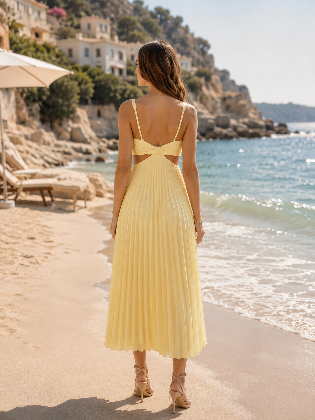 A woman stands on a sandy beach facing the ocean, wearing a pale yellow pleated midi dress with thin spaghetti straps and a crossover back detail. The background features a rocky coastline with buildings and a clear blue sky.