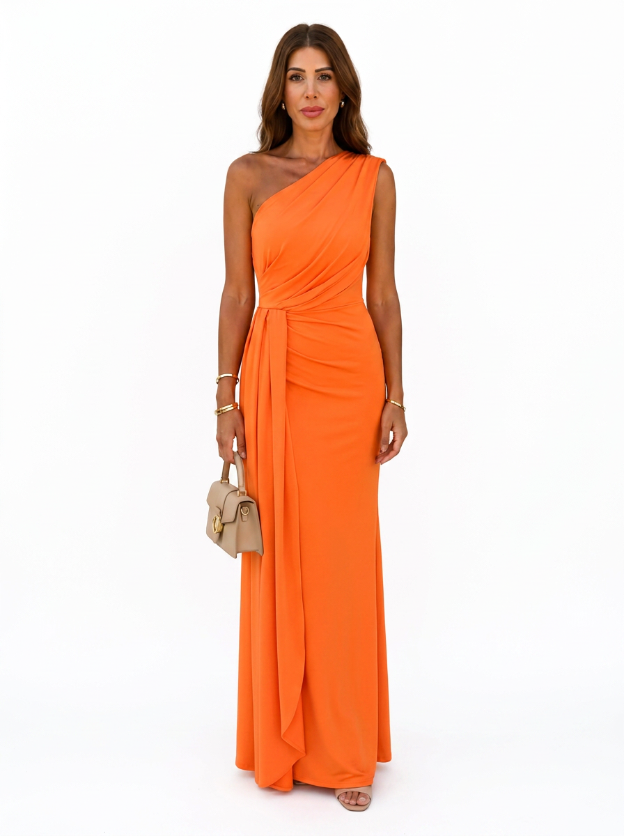 A woman modeling a bright orange one-shoulder floor-length gown with a draped neckline and gathered waist detail, standing against a plain white studio background and holding a small beige handbag.