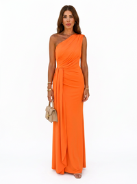 A woman modeling a bright orange one-shoulder floor-length gown with a draped neckline and gathered waist detail, standing against a plain white studio background and holding a small beige handbag.