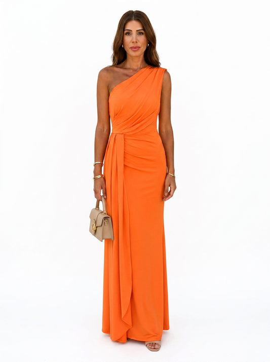A woman modeling a bright orange one-shoulder floor-length gown with a draped neckline and gathered waist detail, standing against a plain white studio background and holding a small beige handbag.