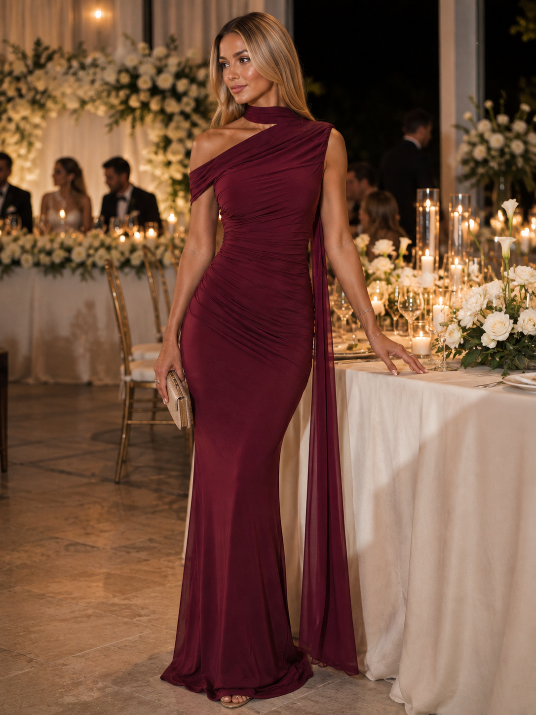 A woman stands at a decorated event table wearing a floor-length, one-shoulder burgundy gown with a draped neckline and side slit. The background features lit tables, floral arrangements, and guests in a warm indoor setting.