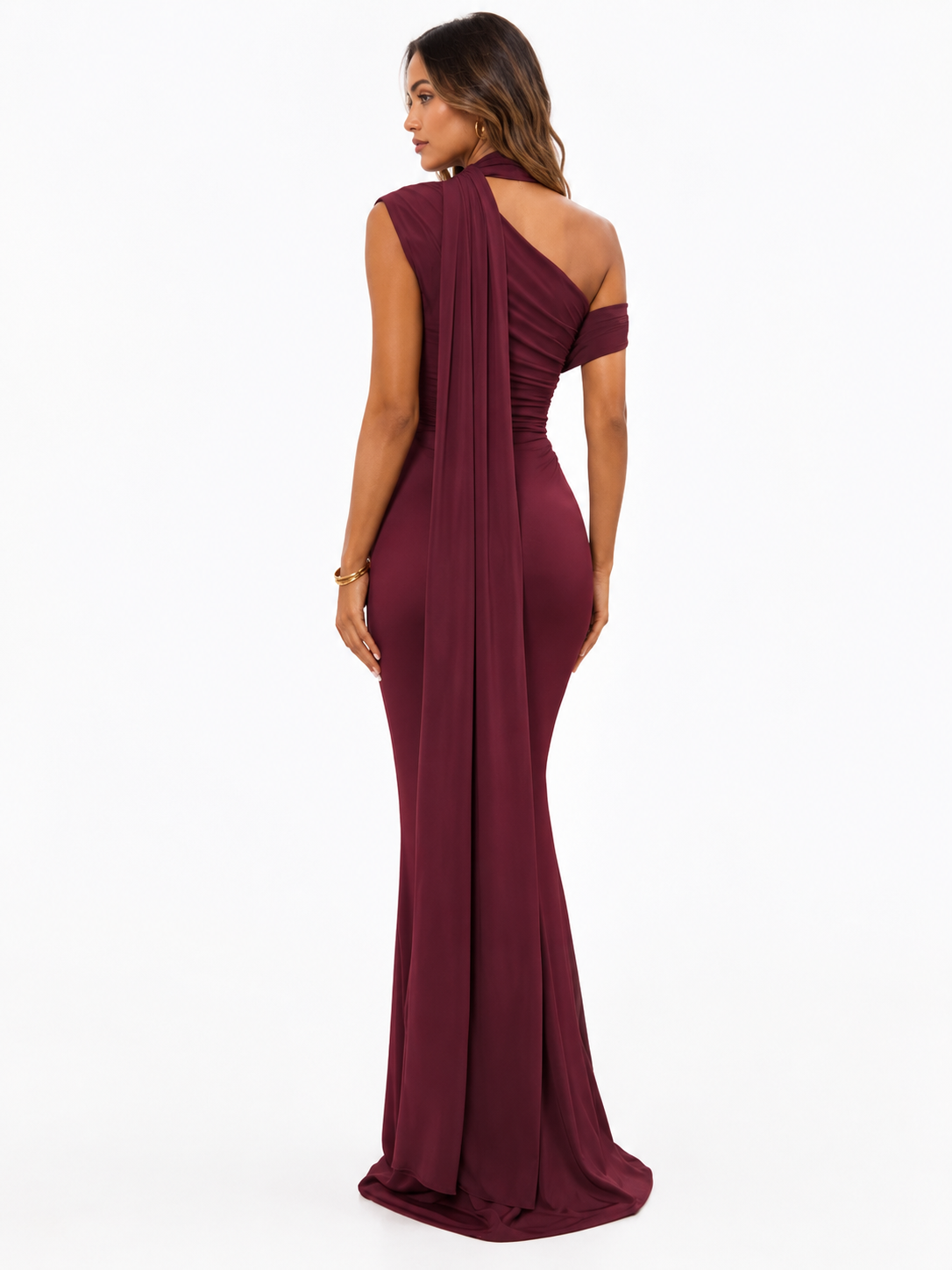 A woman modeling a floor-length, one-shoulder draping gown in deep burgundy from the back, shown against a plain white studio background.